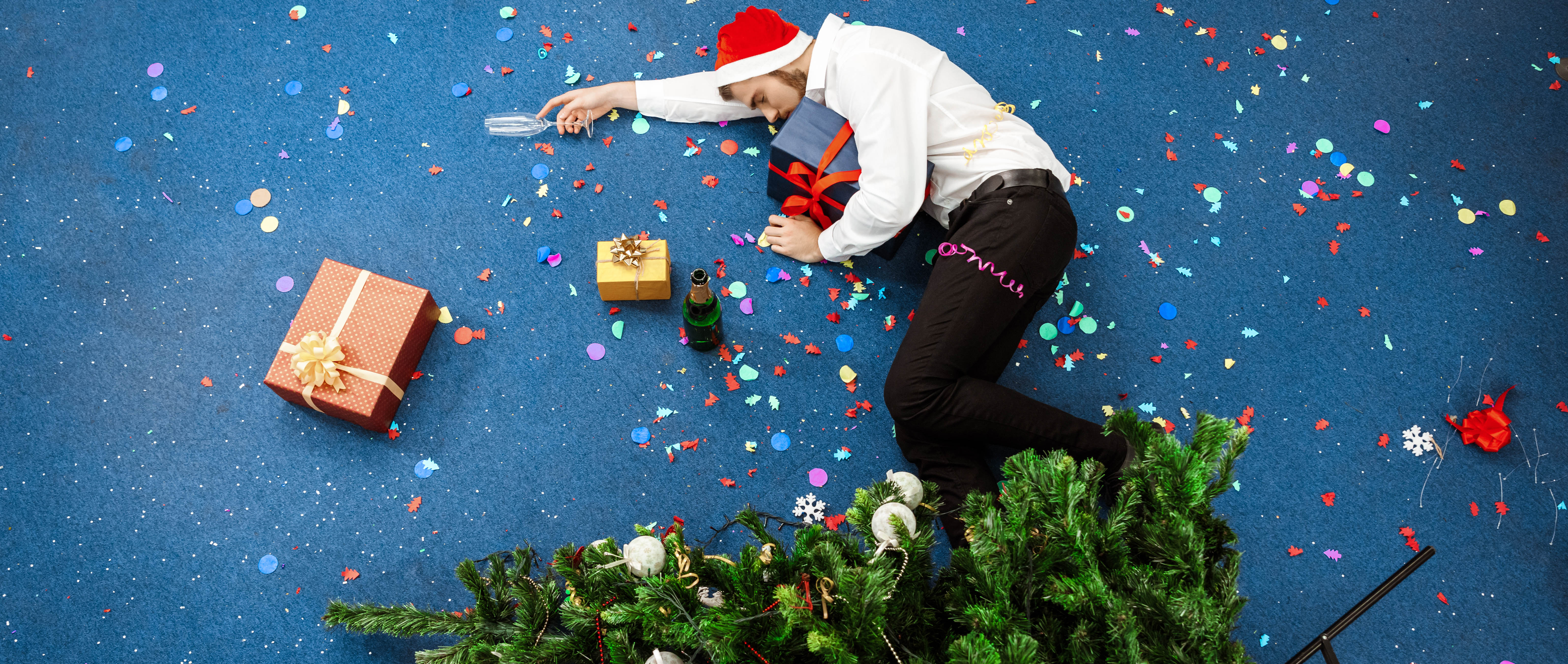 Man asleep on the floor after a holiday party - empty champagne glass in his hand, Santa hat on his head, and Christmas tree tipped down onto his legs